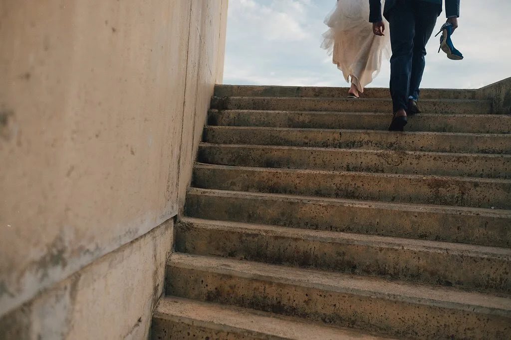 A couple walking up outdoor concrete stairs, the woman in a wedding dress and the man in a dark suit, holding a child’s shoe in his hand.