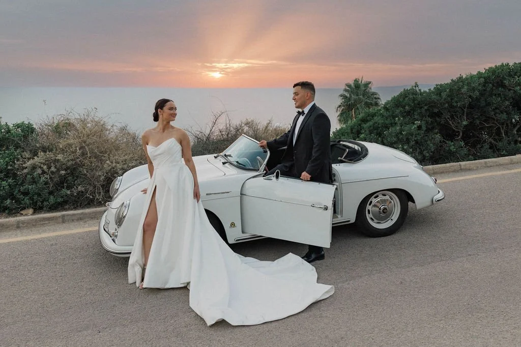 A bride in a white wedding gown standing next to a groom in a black tuxedo, both beside a vintage white convertible car on a scenic roadside during sunset, with greenery and the ocean in the background.