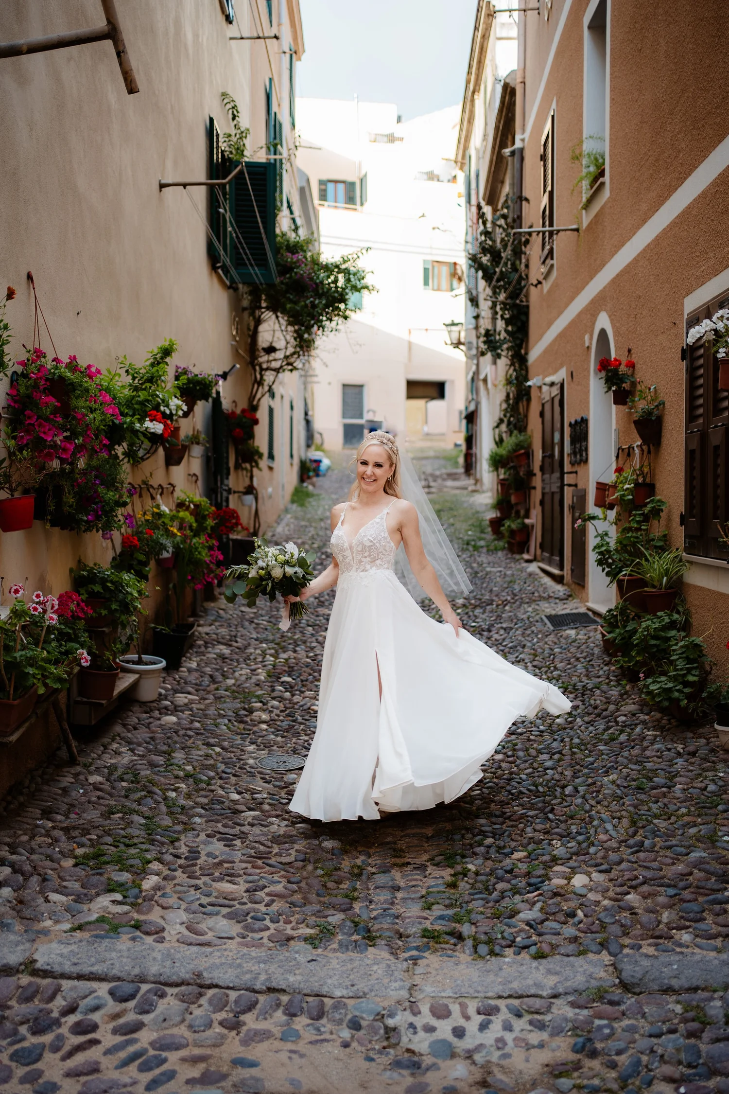 A smiling bride in a white wedding dress holding a bouquet, standing on a cobblestone alley with colorful potted flowers on the walls.