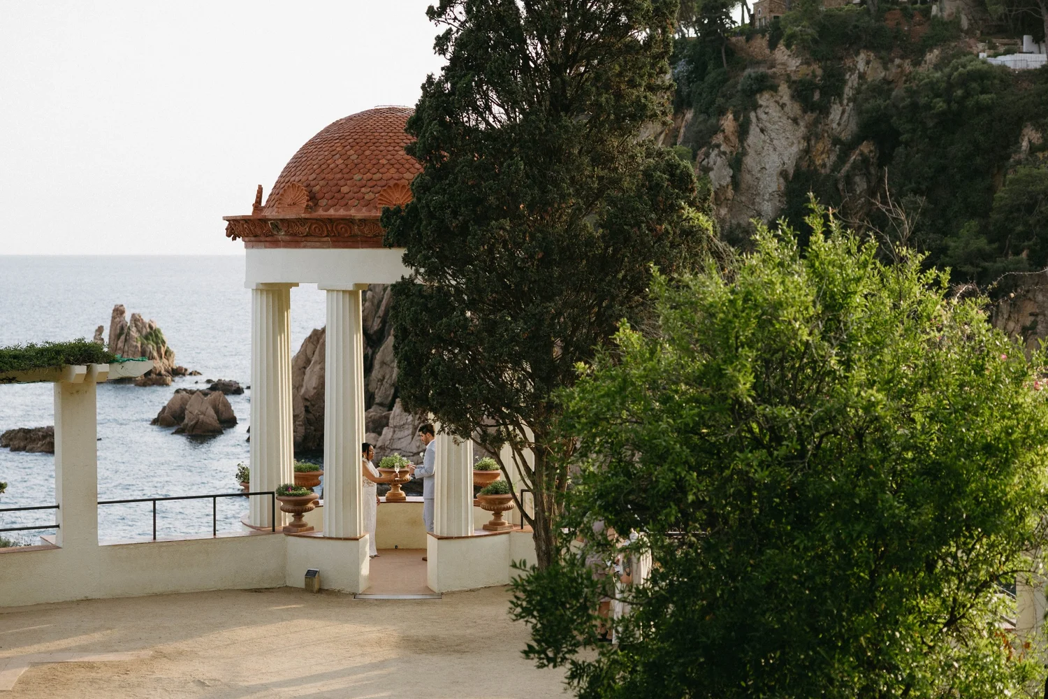 Couple dressed in white dress and suit having a meal at a seaside restaurant with classical architecture, green trees, and rocky cliffs in the background.