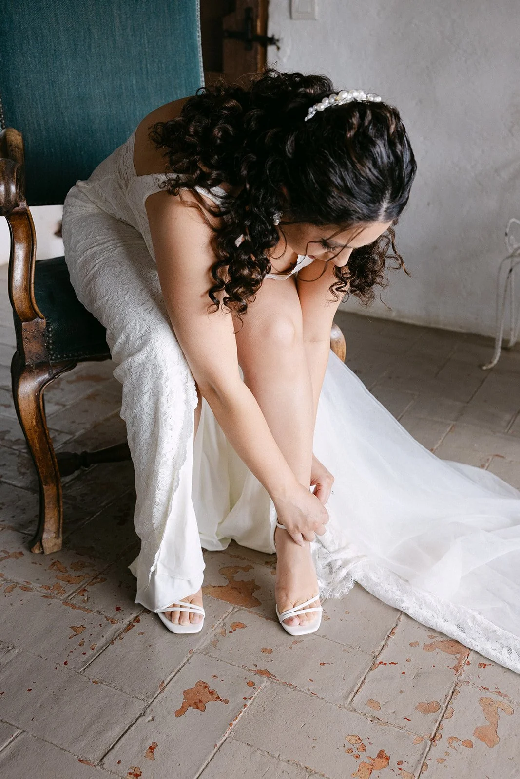 A bride with curly hair and a pearl hairpiece is sitting on a vintage wooden chair, adjusting her white high-heeled shoe while wearing a white wedding dress.