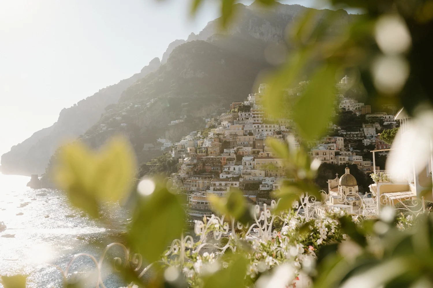 A hillside town with white and pastel-colored buildings overlooking the ocean, with some flesh-colored and white domed structures in the foreground, partly obscured by blurred greenery.