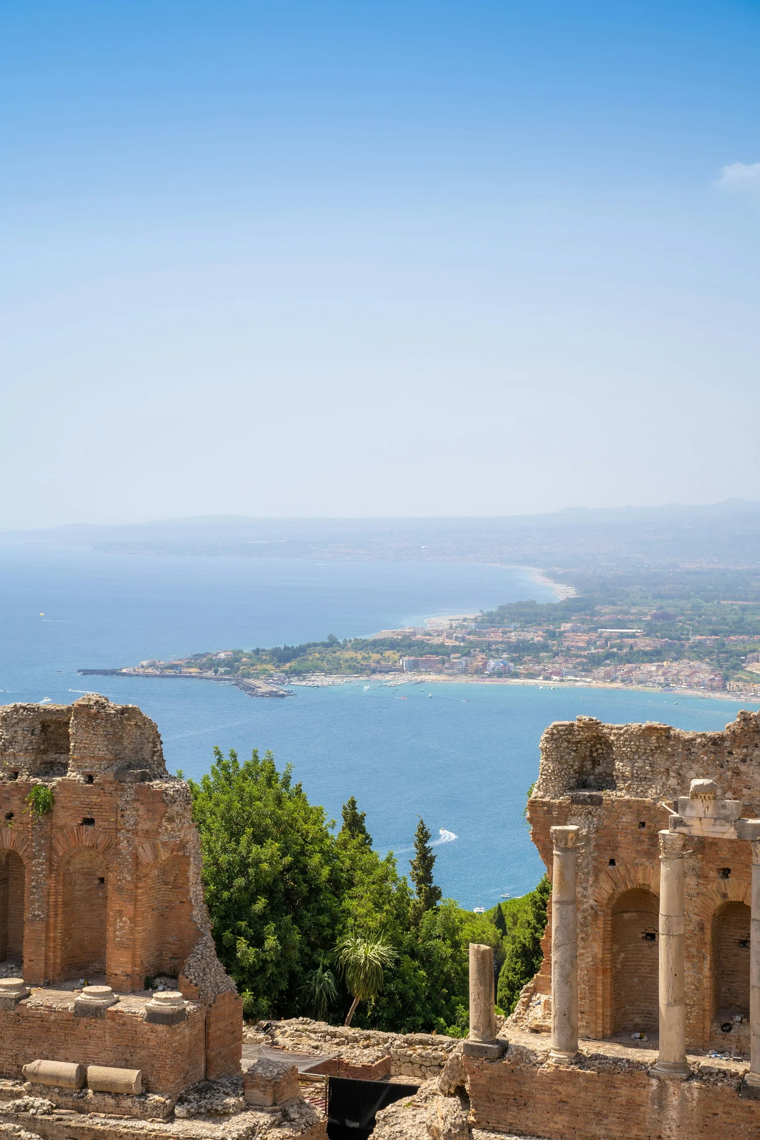 Ancient ruins with brick and stone walls and columns, overlooking a coastline with blue water and clear sky.