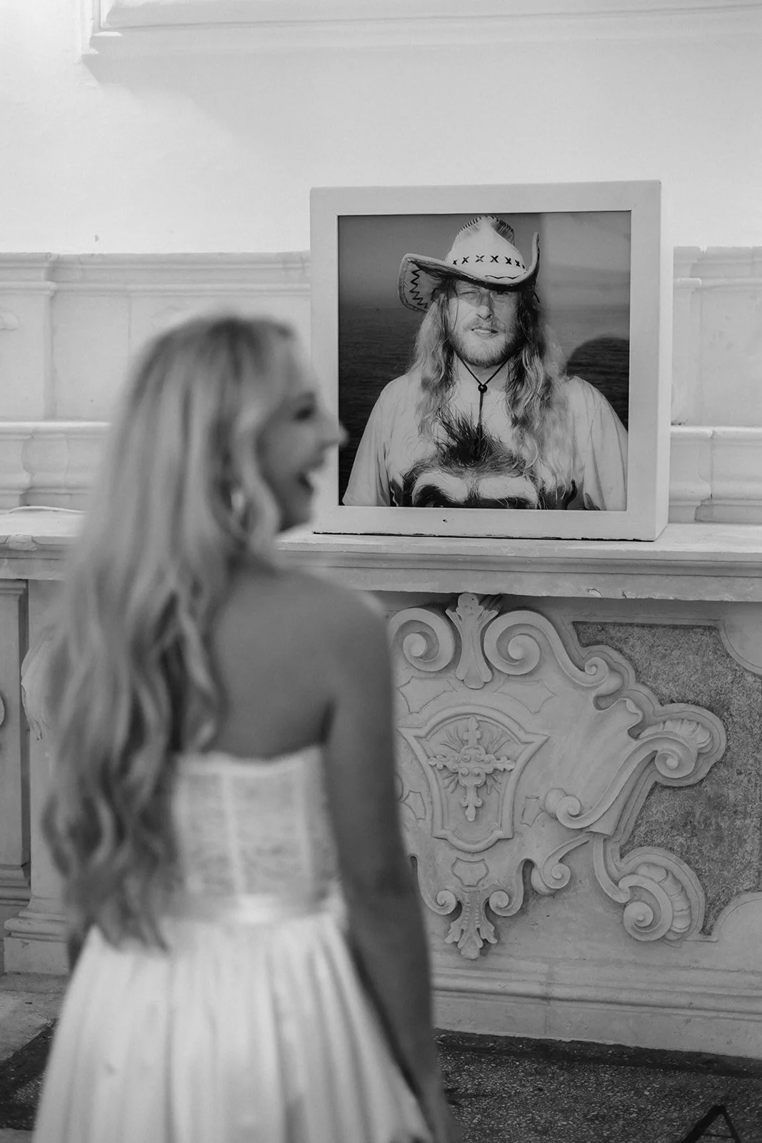 A woman with long hair in a strapless dress looking at a framed photograph of a man in a cowboy hat on a mantle in an indoor setting.