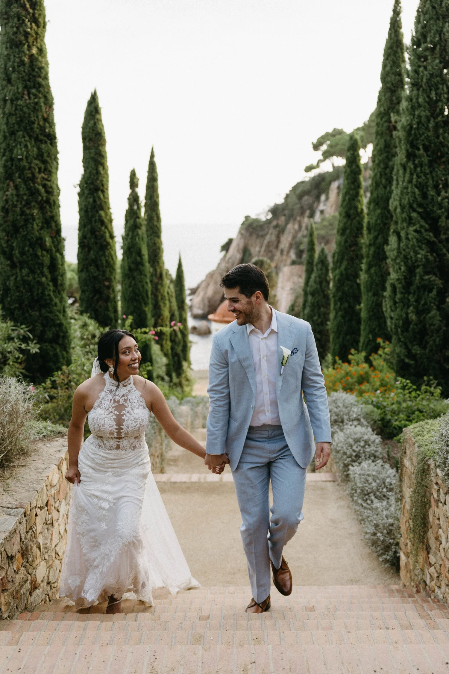 A bride and groom walking hand in hand down a garden path, smiling at each other, surrounded by tall green cypress trees and colorful flowers.