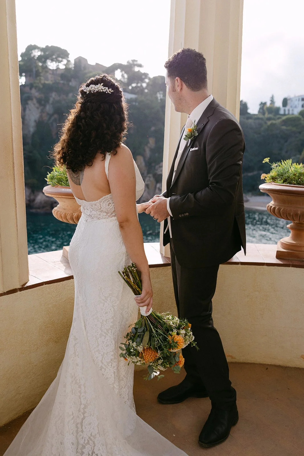 A bride and groom exchanging vows at a wedding ceremony, overlooking a scenic water and hillside view, with the bride holding a bouquet of flowers.