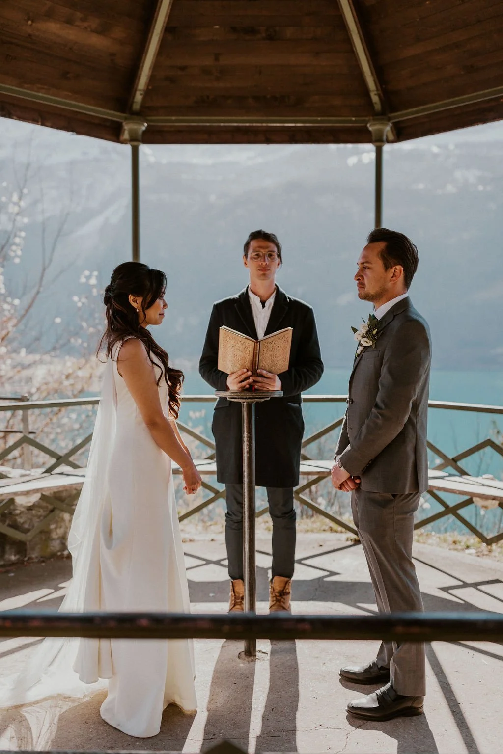 A bride and groom facing each other during their wedding ceremony outdoors, with an officiant standing behind a stand with a book, in front of a scenic mountain landscape.