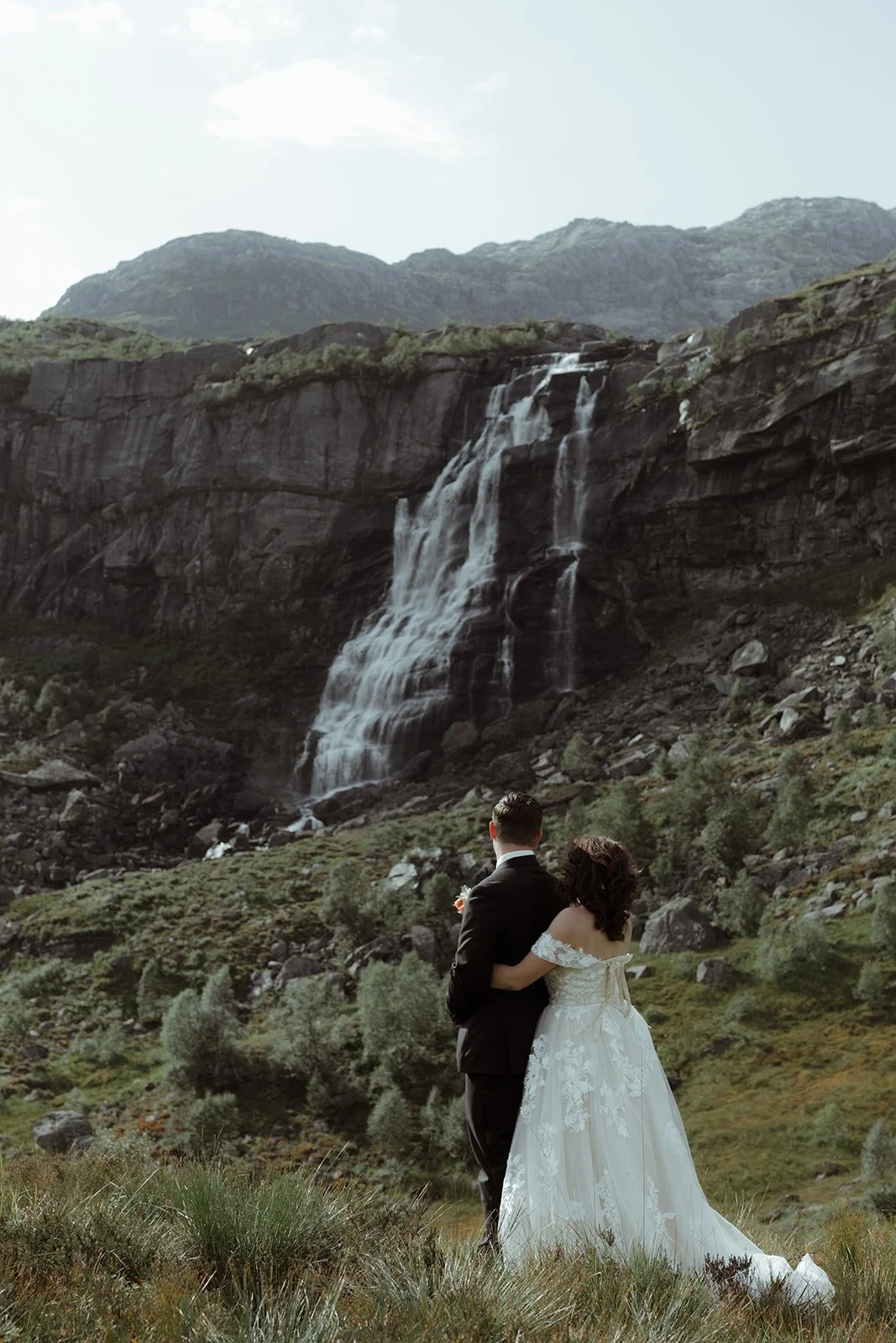 A couple dressed in wedding attire standing in a grassy area with a large waterfall and mountain in the background.