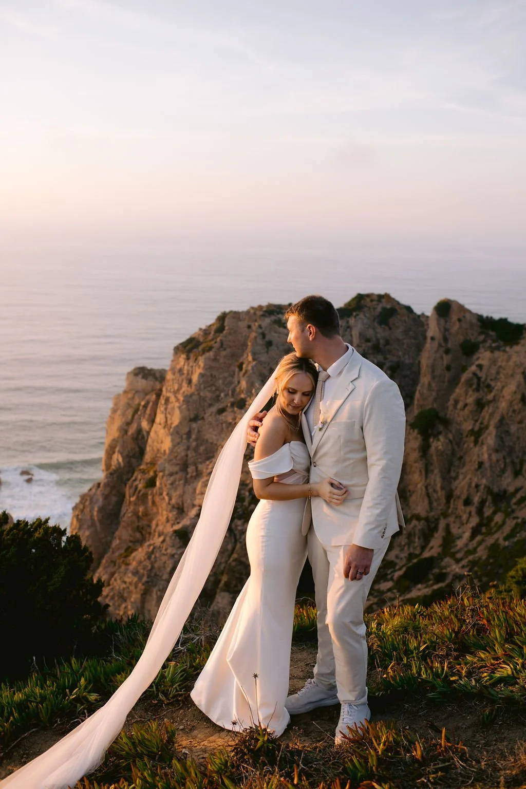 A bride and groom dressed in white embrace on a cliffside overlooking the ocean at sunset.