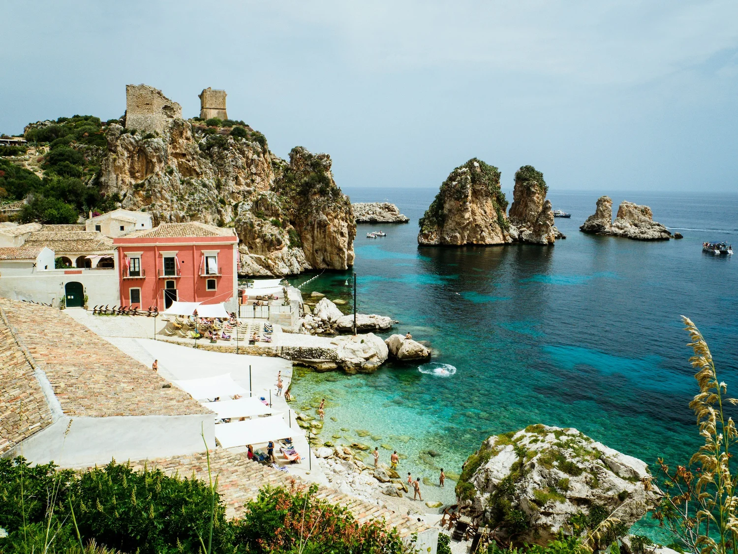 Cliffs with ruins on top overlooking the sea, colorful buildings at the base, people swimming and relaxing at the beach, boats on calm waters.