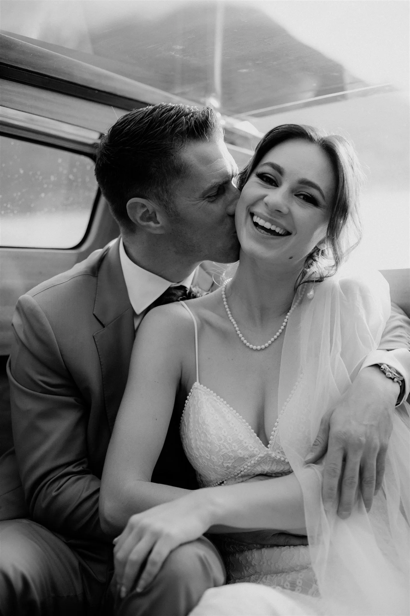 A black-and-white photo of a couple, a man and woman, sitting close together, with the man kissing the woman's cheek. The woman is smiling, wearing a lace dress and pearl necklace. The man is dressed in a suit and tie. They are inside a vehicle with