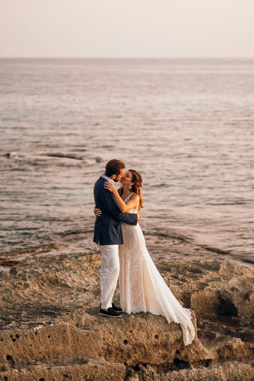 A couple dressed in wedding attire sharing a kiss on a rocky shoreline with the ocean in the background at sunset.