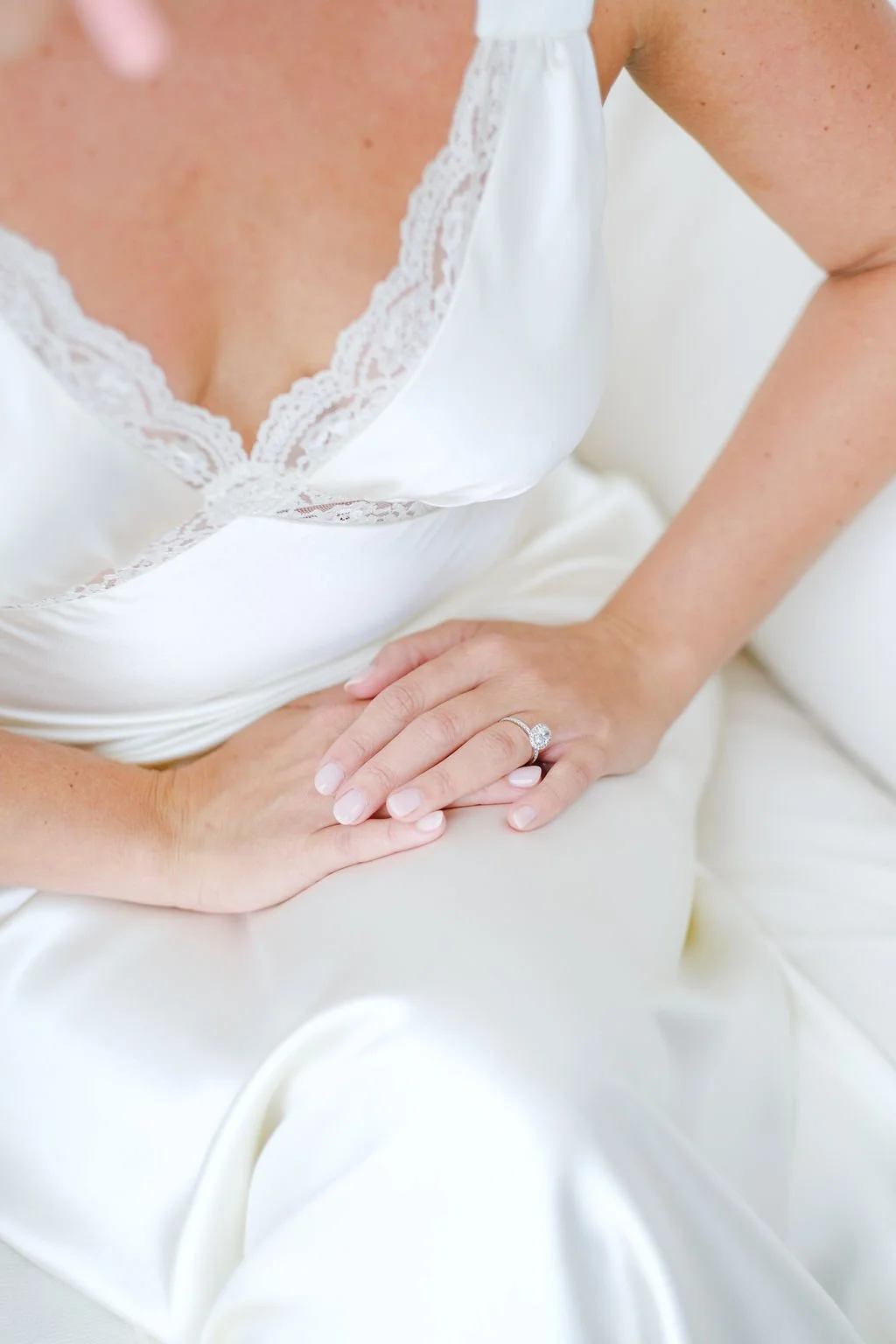 Close-up of a woman in a white satin nightgown with lace trim, wearing an engagement ring on her left hand, resting her hand on her thigh.