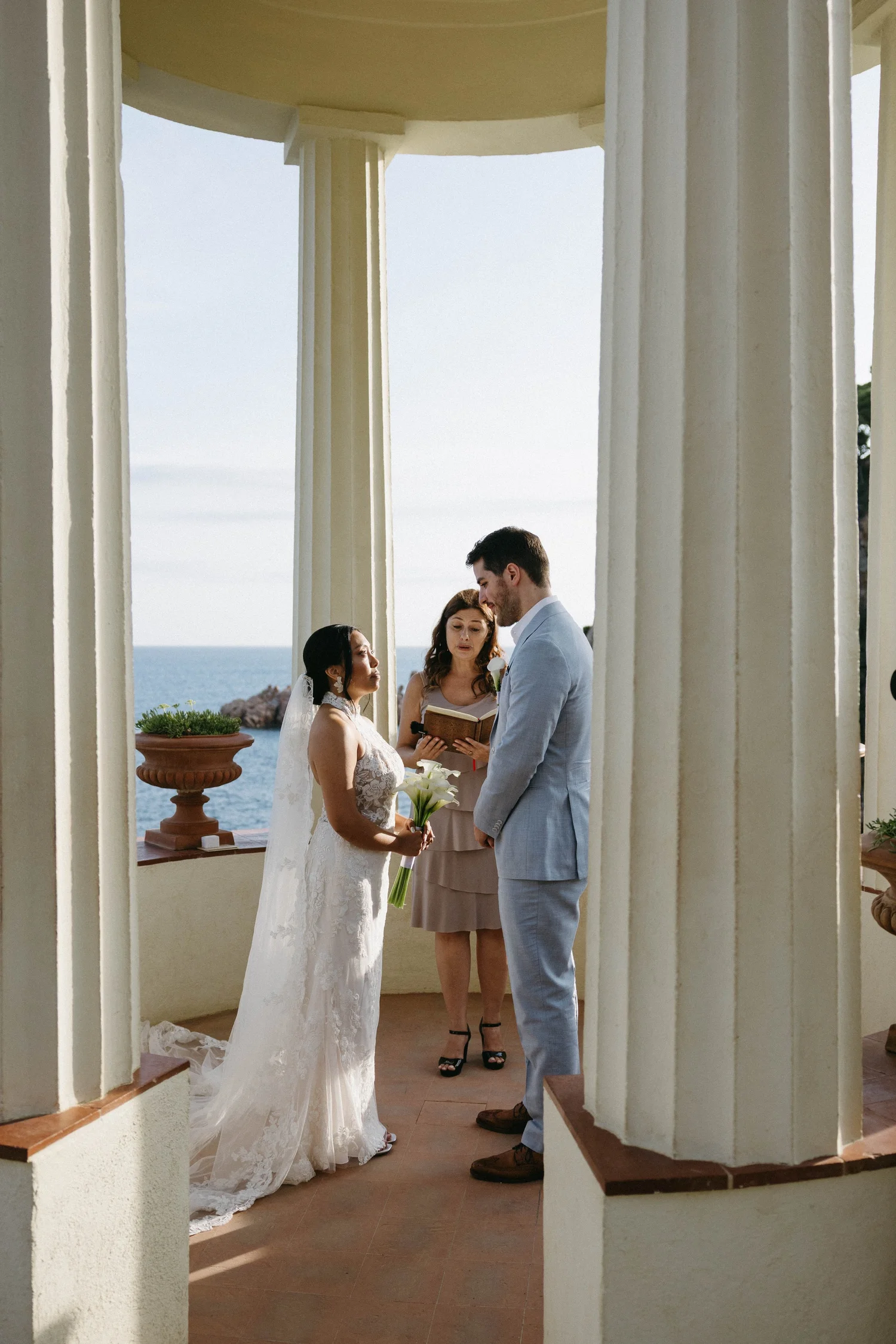A wedding ceremony taking place outdoors on a balcony with ocean view in the background. A bride in a lace wedding dress and a groom in a light-colored suit are facing each other, holding hands, while a woman officiant reads from a book.