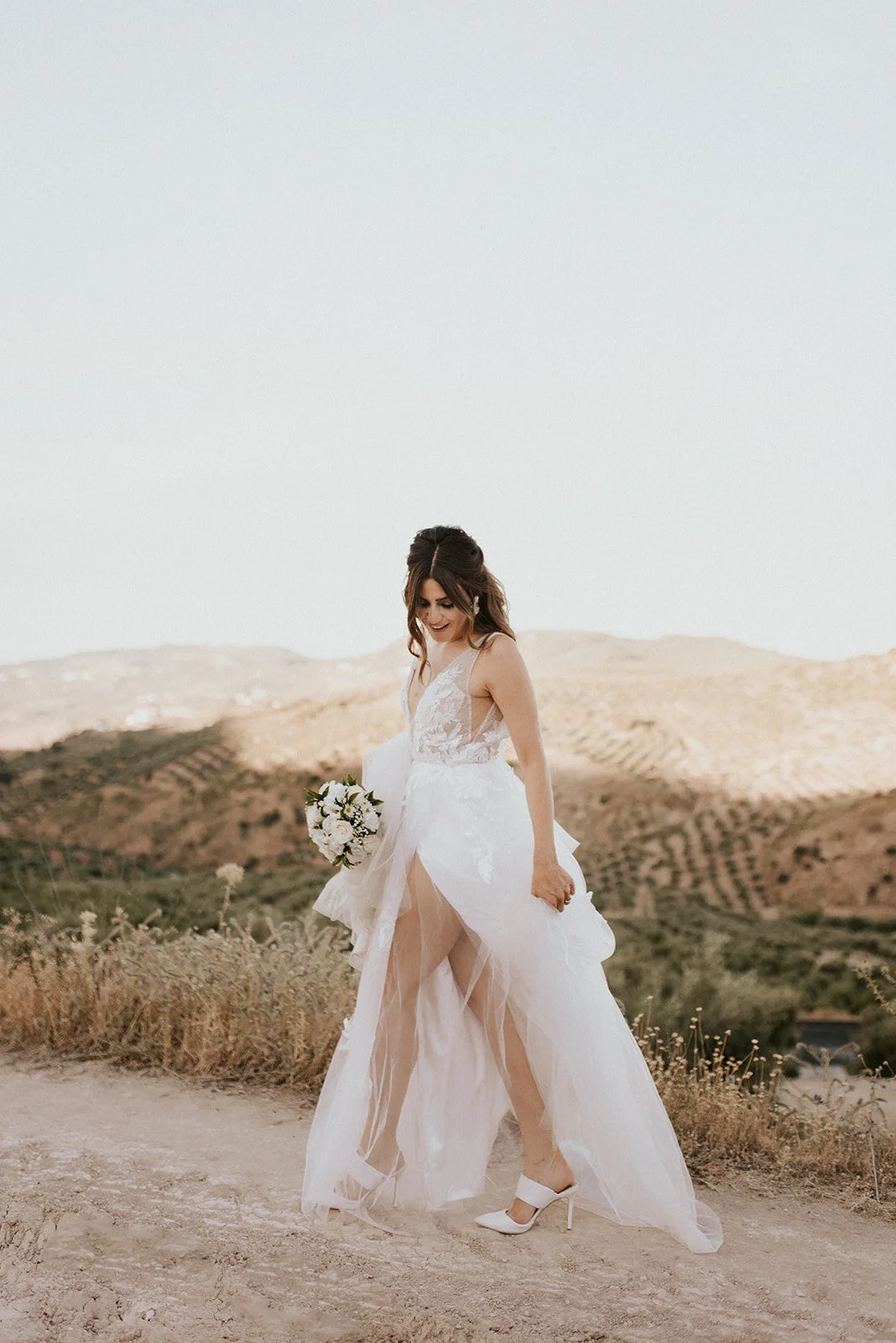 A bride in a white wedding dress holding a bouquet, standing outdoors on a dirt path with hills in the background during daytime.