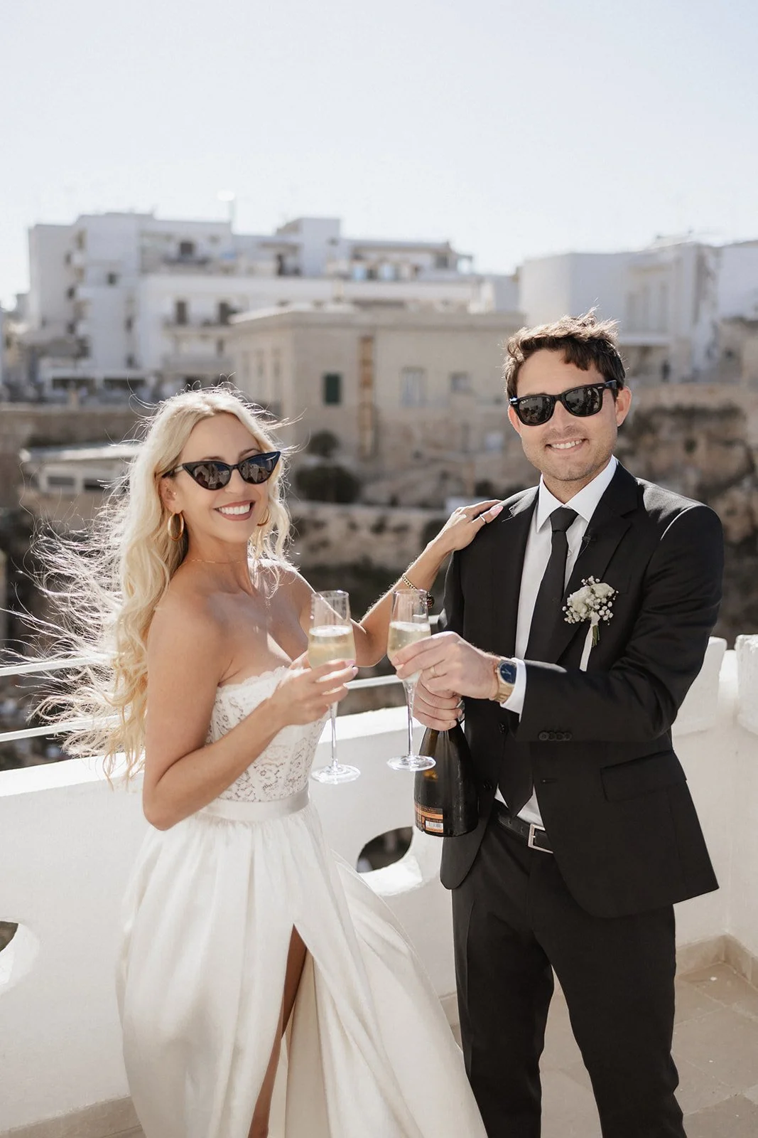 A bride and groom celebrating with champagne on a balcony with a white building and bright sky in the background.