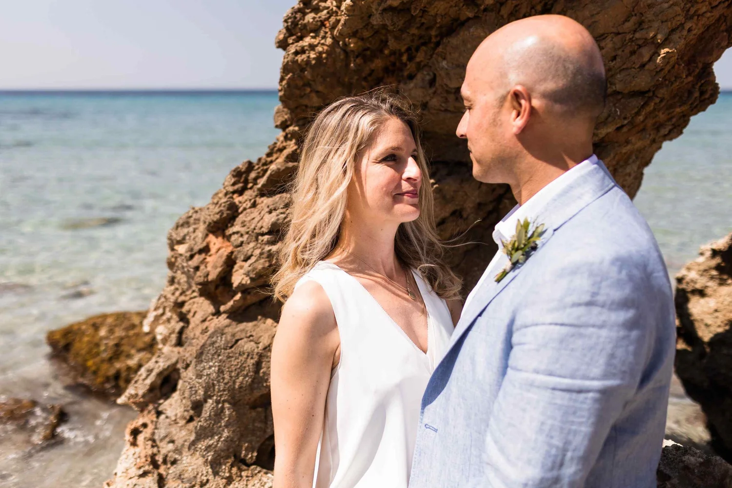 A couple standing close together on a beach with large rocks behind them, facing each other and smiling.