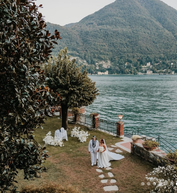 A couple in wedding attire walking on a stone pathway near a lake with mountains in the background, decorated with white flowers and outdoor wedding setup.
