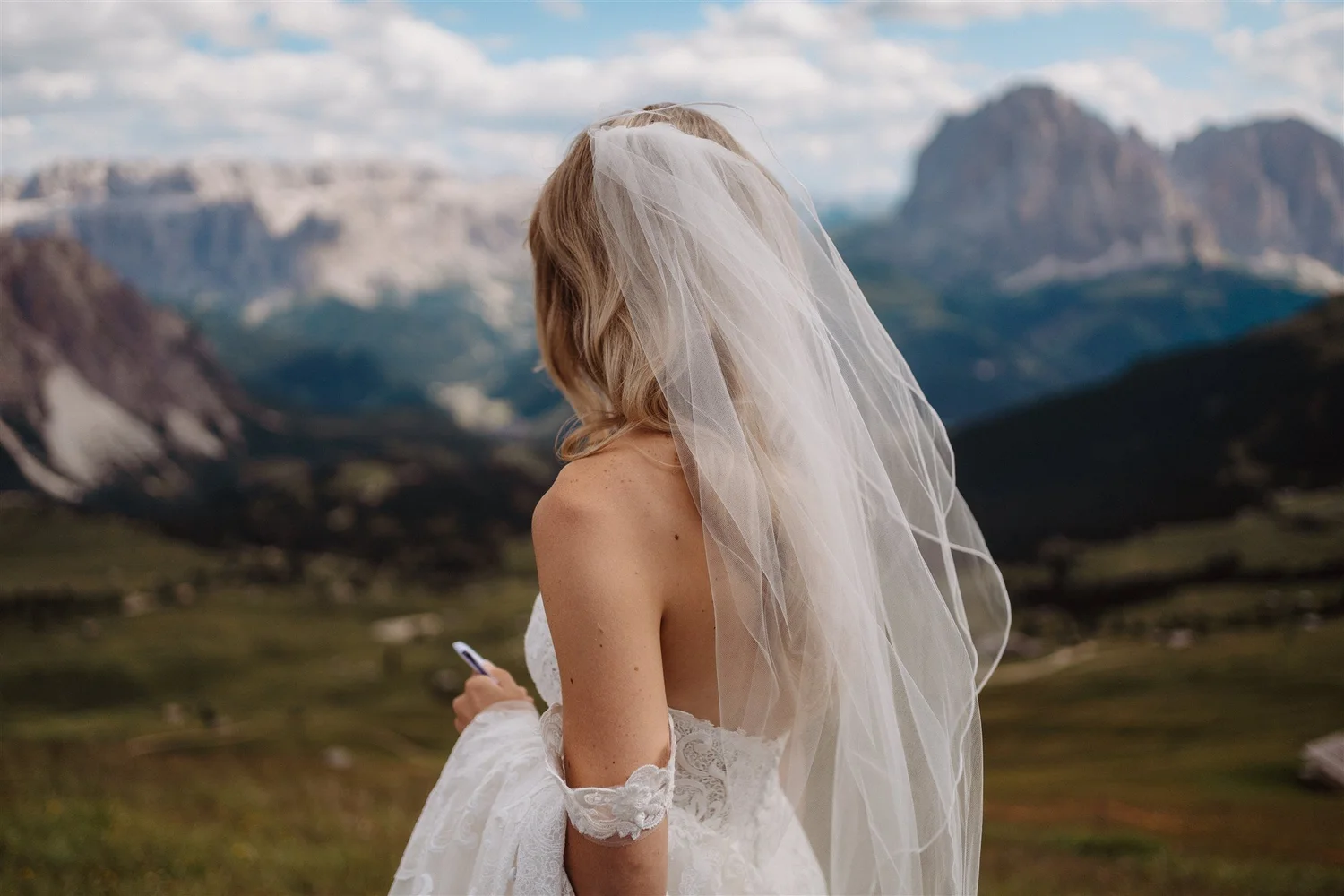A woman in a wedding dress with a veil is outdoors in front of mountains under a partly cloudy sky.