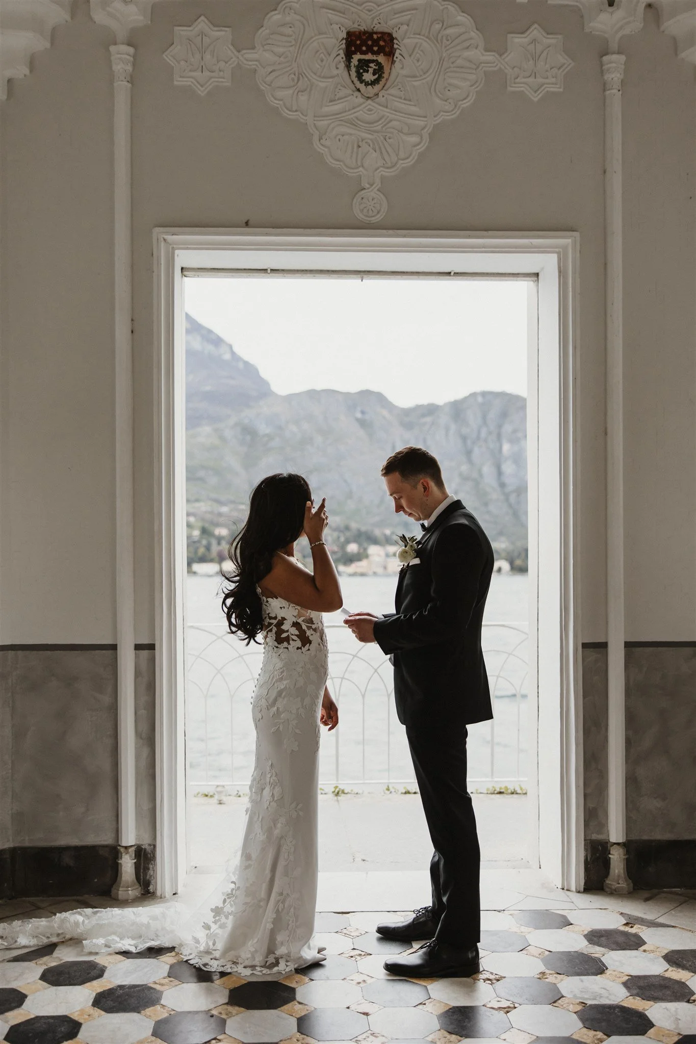 A bride and groom standing face to face in front of a large window with mountains in the background, holding hands, during their wedding ceremony.
