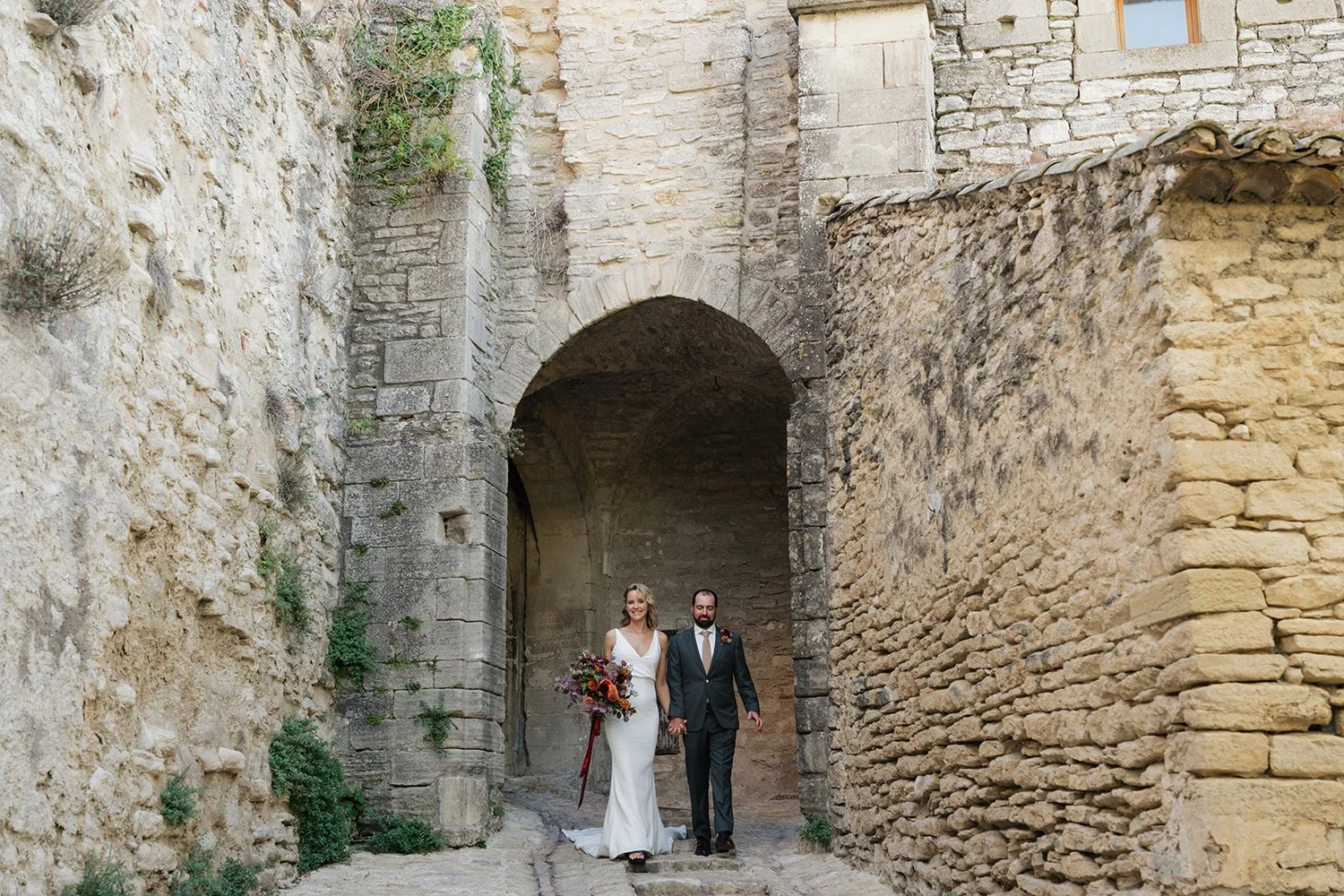 A bride and groom walking hand in hand through an ancient stone archway, with rustic stone walls and greenery around them.