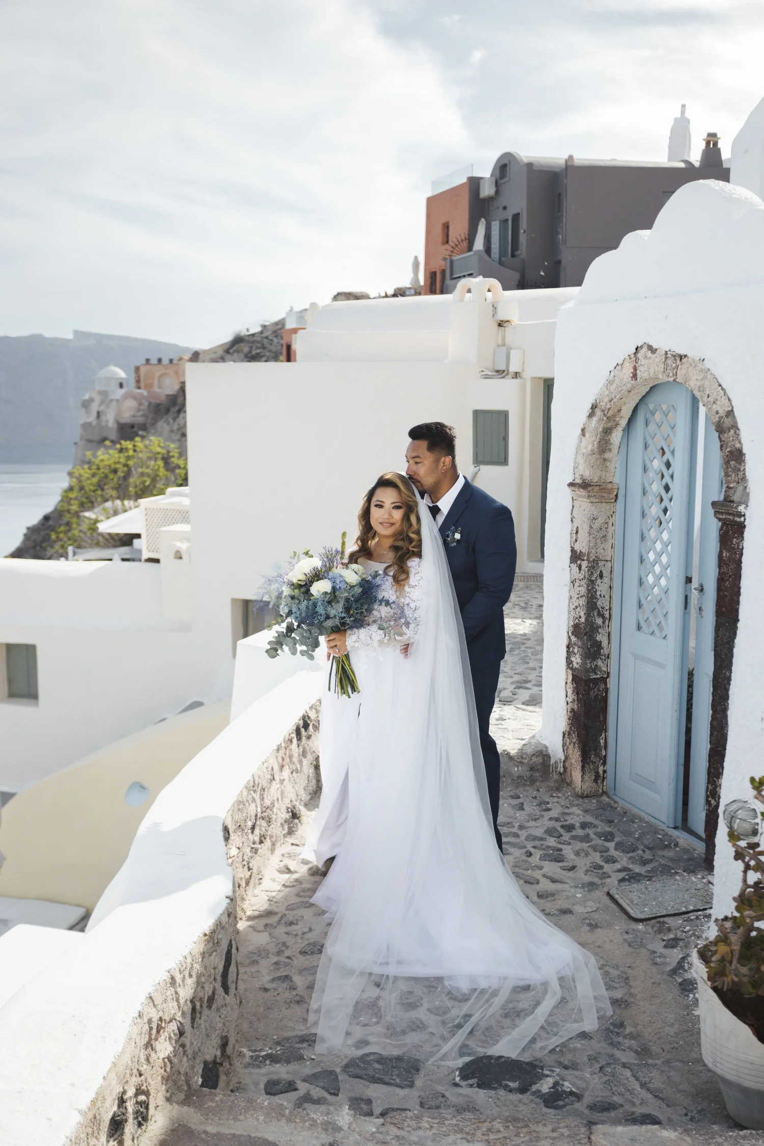 A bride in a white wedding dress holding a bouquet, standing next to a groom in a navy suit, with white buildings and a blue door in the background, on a stone pathway.