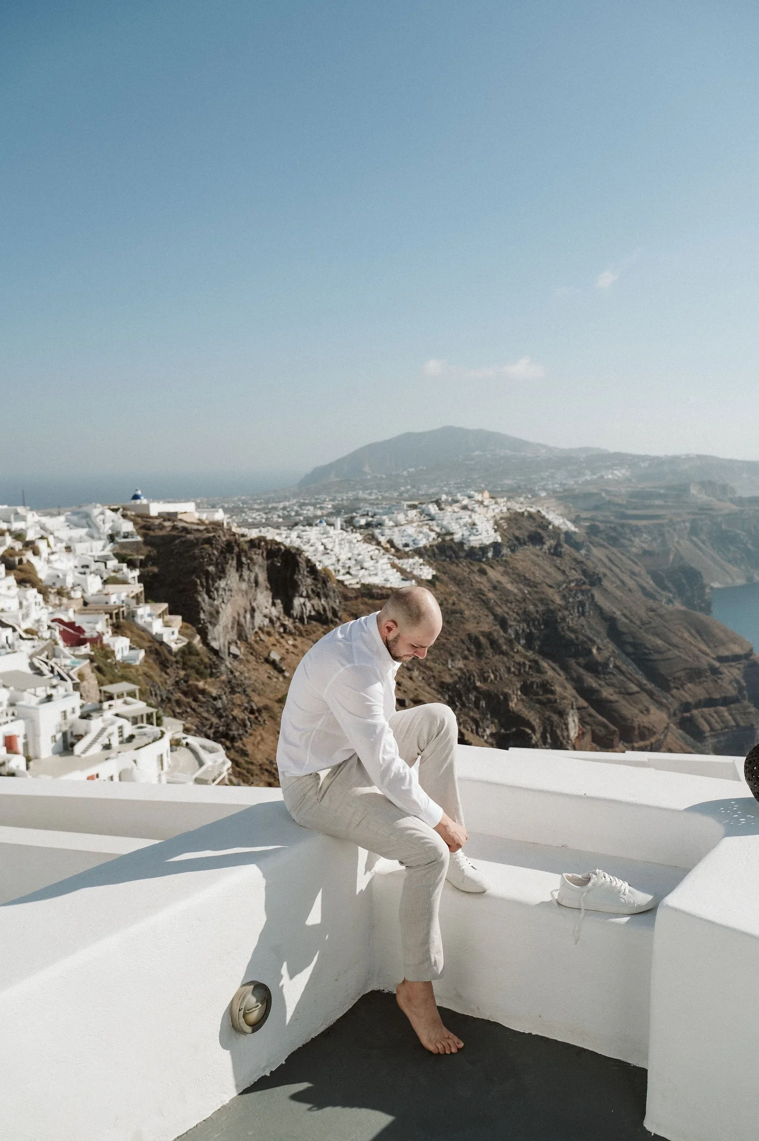 A man dressed in white, sitting on a white ledge, tying his shoelaces against a backdrop of white buildings and a hillside landscape with a mountain in the distance under a clear sky.