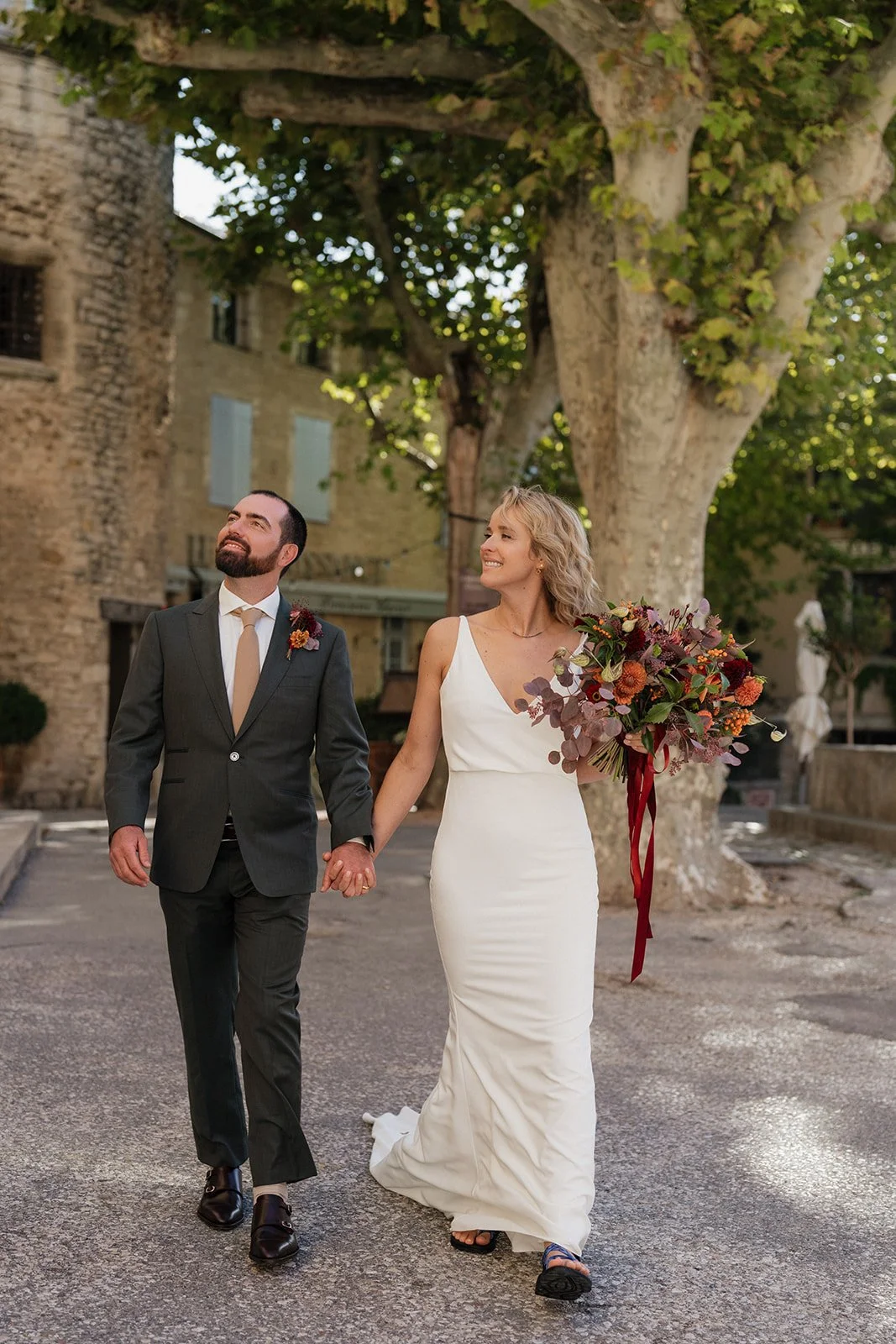 A newlywed couple walking hand in hand outdoors; the woman is holding a large bouquet of flowers and wearing a white wedding dress, the man is in a dark suit with a beige tie. They are walking on a street lined with trees and old buildings, looking h