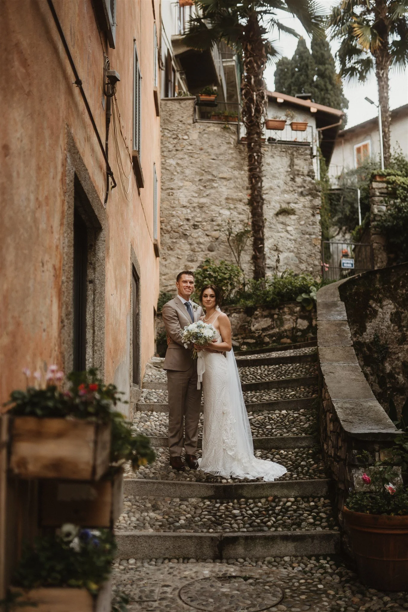 A bride and groom standing in a narrow alleyway with aged walls, cobblestone steps, potted plants, and palm trees, dressed in wedding attire, smiling at the camera.