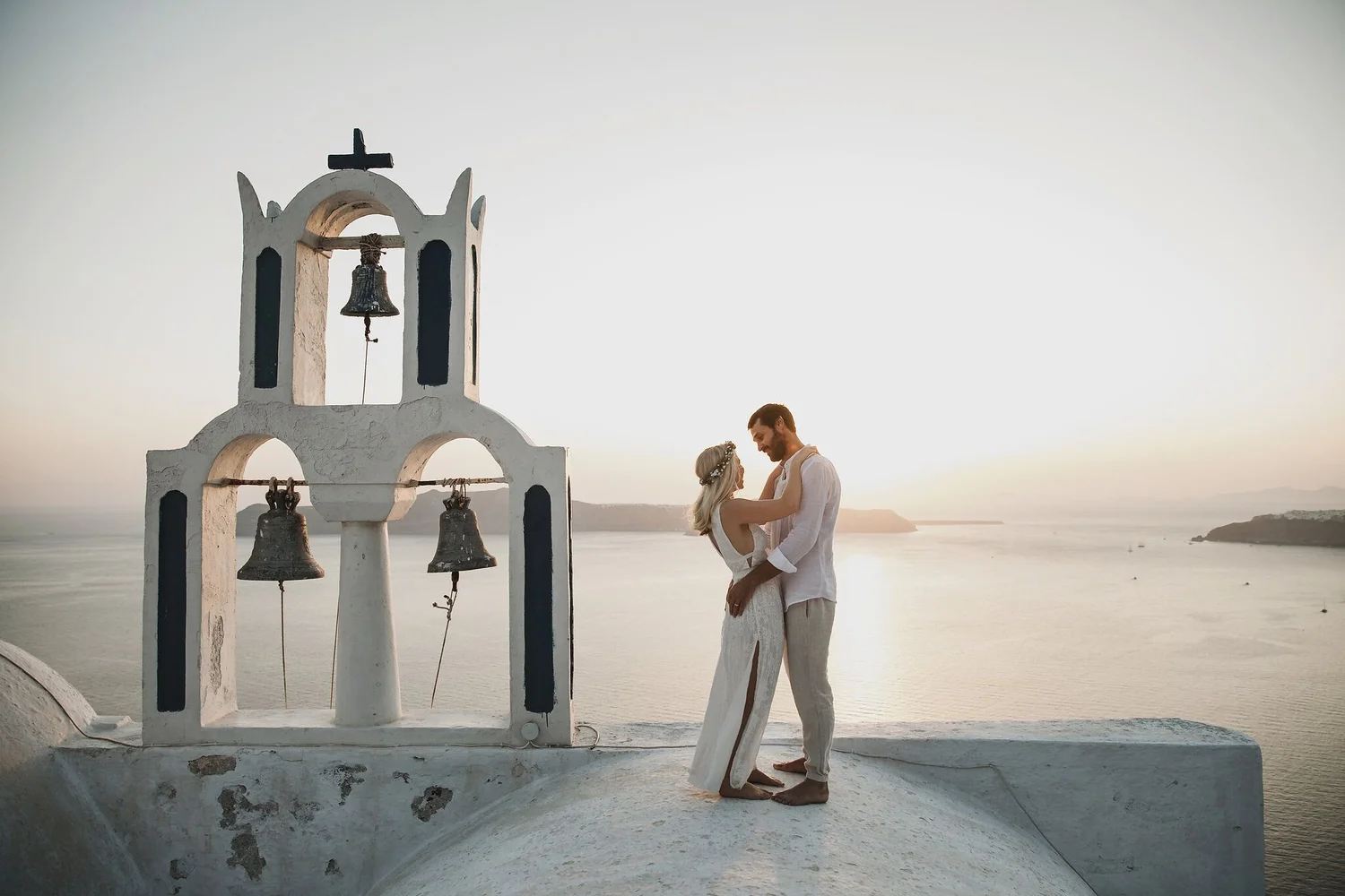 A couple dressed in white, standing close together on a white rooftop with a scenic ocean view and cliffs in the background at sunset.