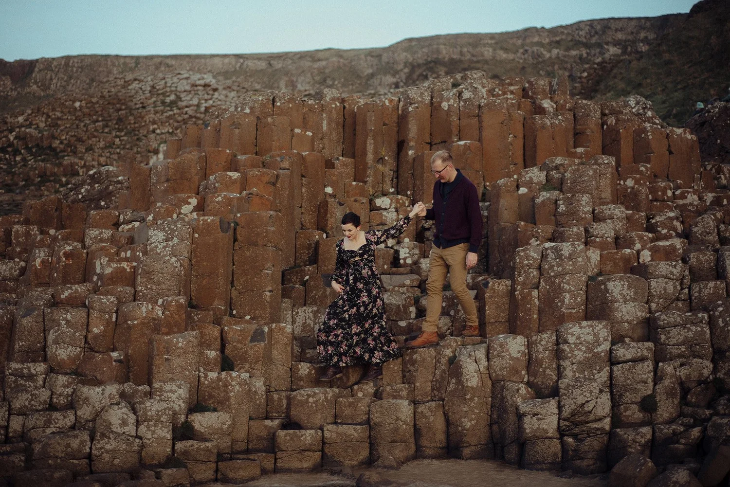A man and a woman holding hands on ancient basalt columns in a landscape, with the woman in a floral dress and the man in a dark jacket and tan pants.