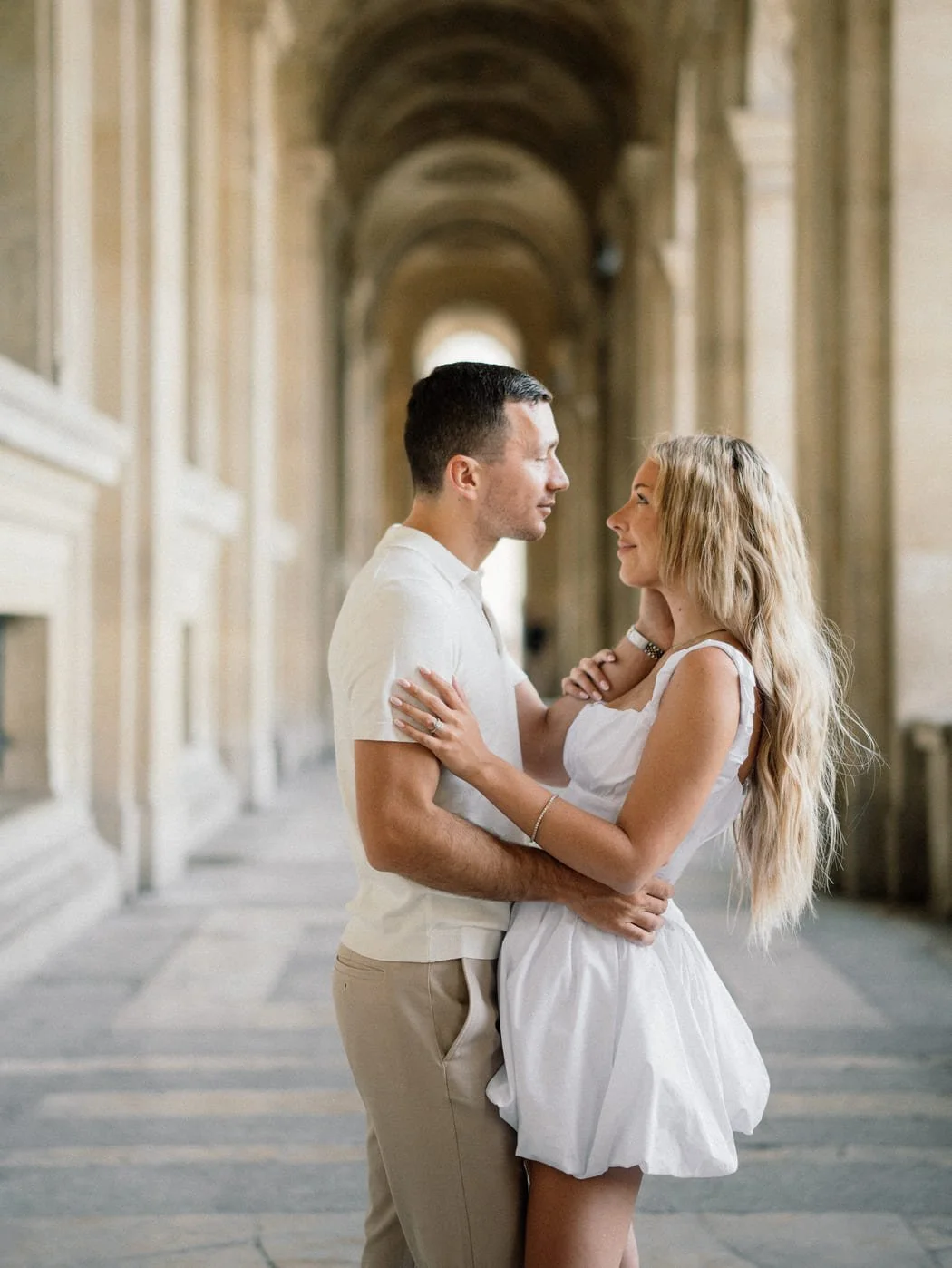 A couple holding each other and gazing into each other's eyes in a grand hallway with arches.