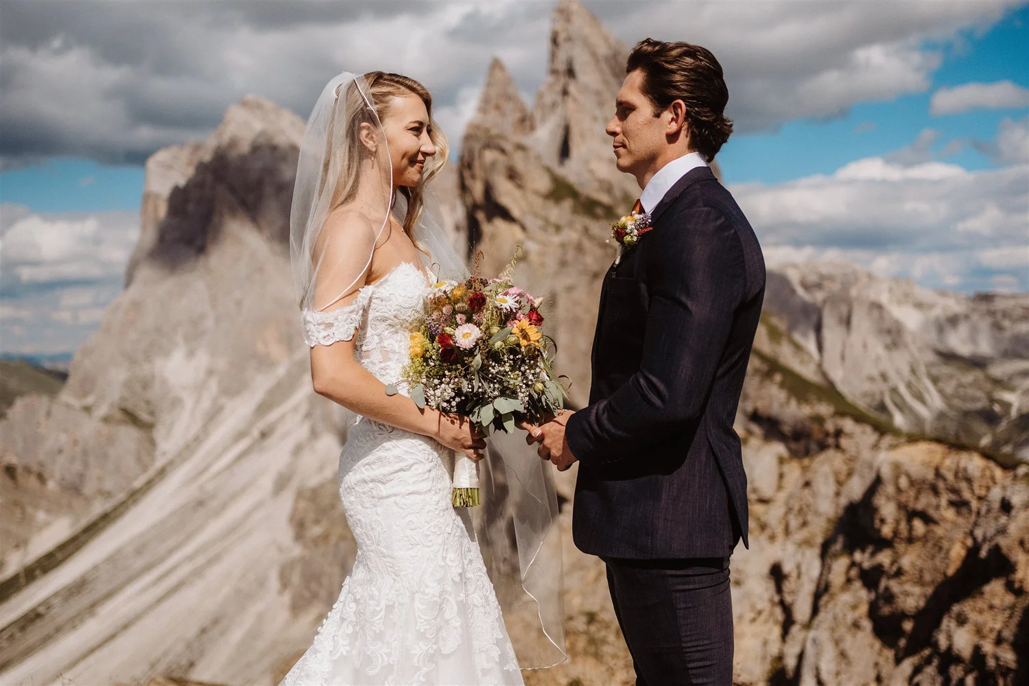 A bride and groom facing each other and holding hands outdoors with rocky mountains in the background.