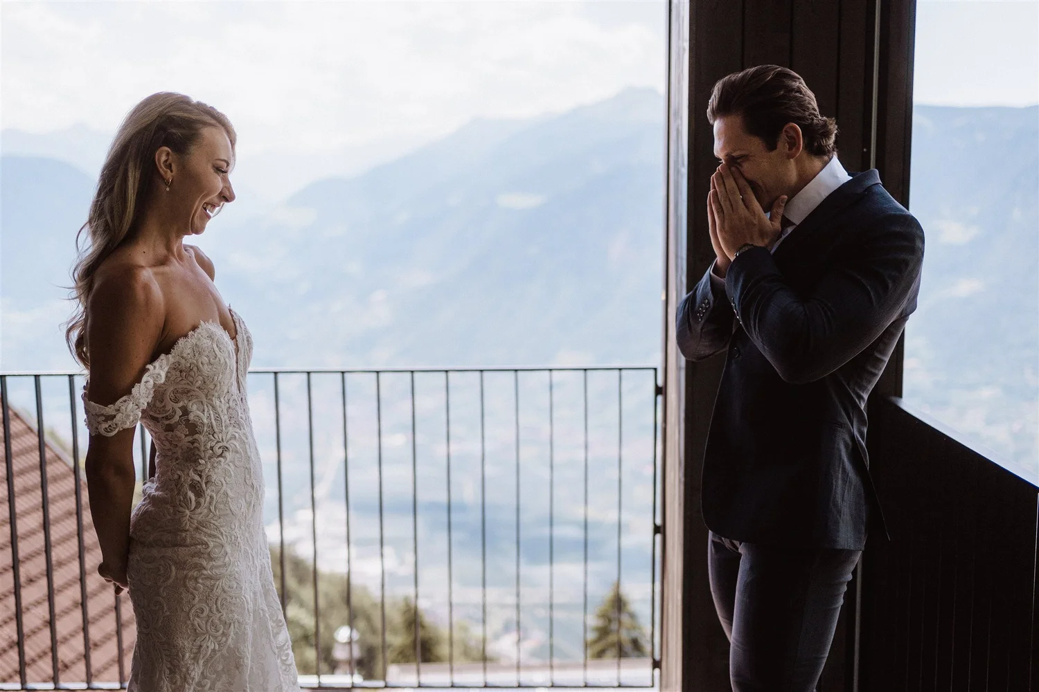 A bride standing in a wedding dress facing a groom in a suit, both smiling and sharing a joyful moment indoors with mountain views in the background.
