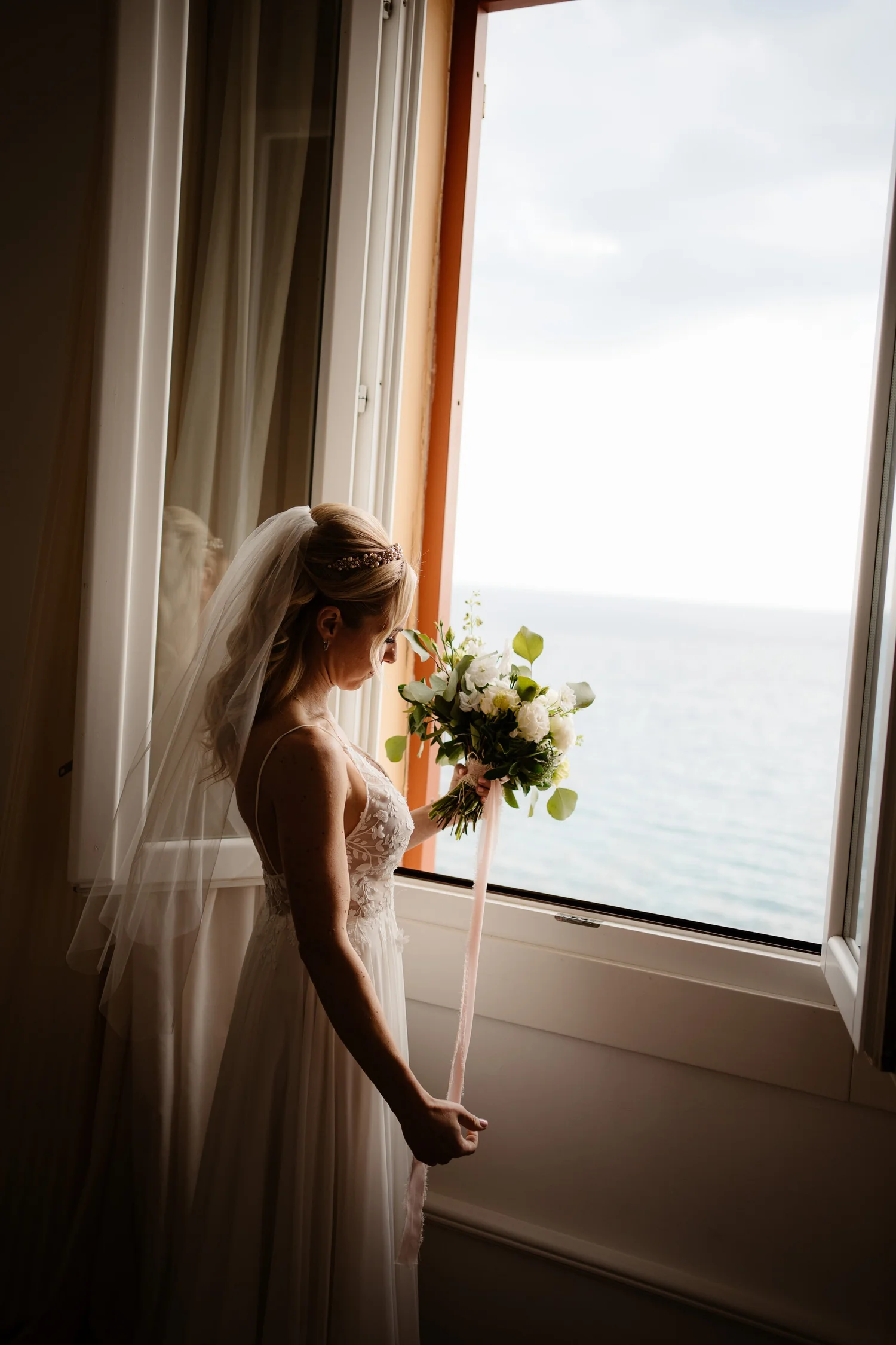 A bride in a white wedding dress holding a bouquet of white and green flowers, standing near an open window with a view of the sky and water, looking down.