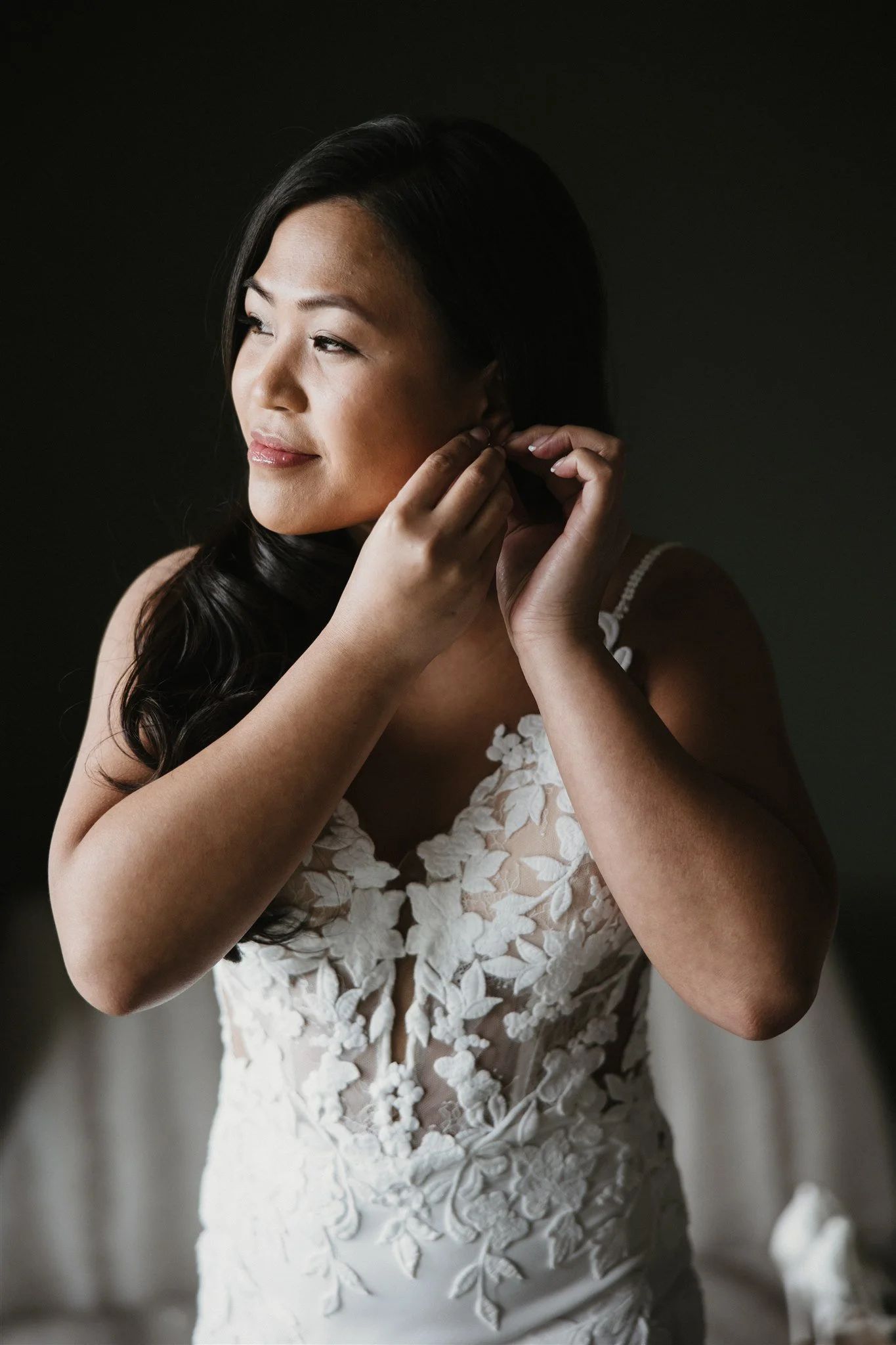 A woman in a white lace dress is putting on earrings. She has long dark hair and a subtle smile, standing against a dark background.