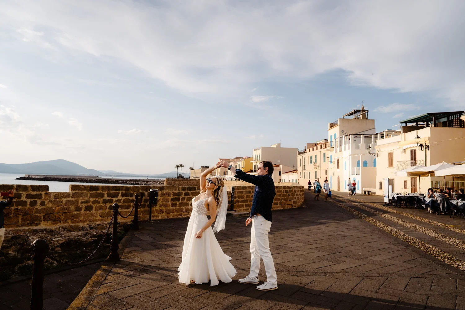 A bride in a white wedding dress and a groom in a white shirt and dark blazer are taking photos together on a seaside promenade with pastel-colored buildings, a cloudy sky, and people relaxing nearby.