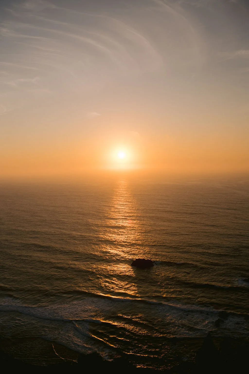 Sunset view over the ocean with a gentle reflection on the water and a single rock in the foreground.