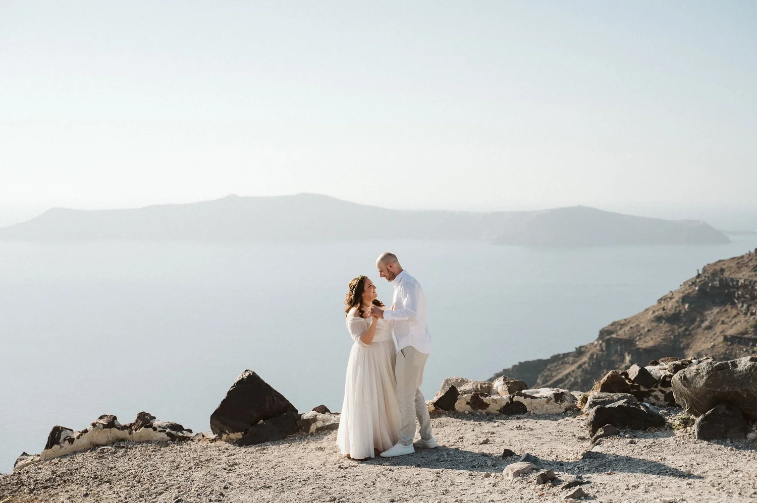 A couple with wedding attire standing close together on a rocky coastline with water and mountains in the background.