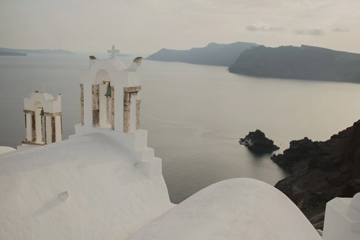 White church with bell towers and crosses overlooking a body of water with mountains in the distance on a cloudy day.