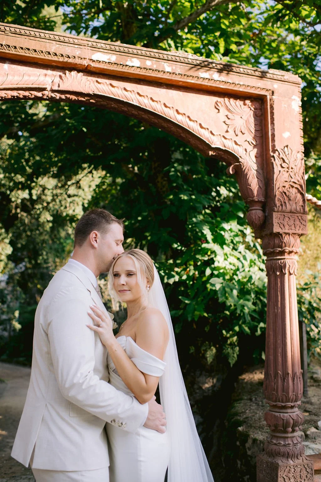 A bride and groom embracing under an ornate wooden arch with lush green foliage in the background.