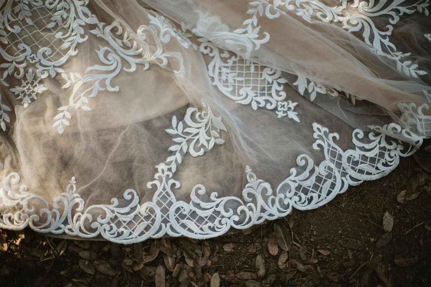 Close-up of delicate lace fabric with floral and grid patterns, draped over ground covered with dry leaves and dirt.