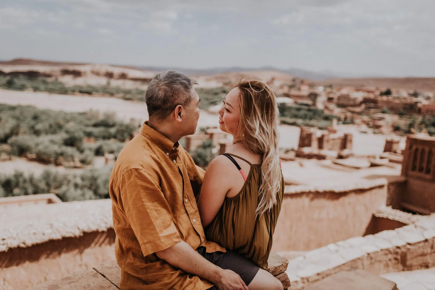 A man and woman sitting closely together on a stone ledge, facing each other with a city landscape in the background.