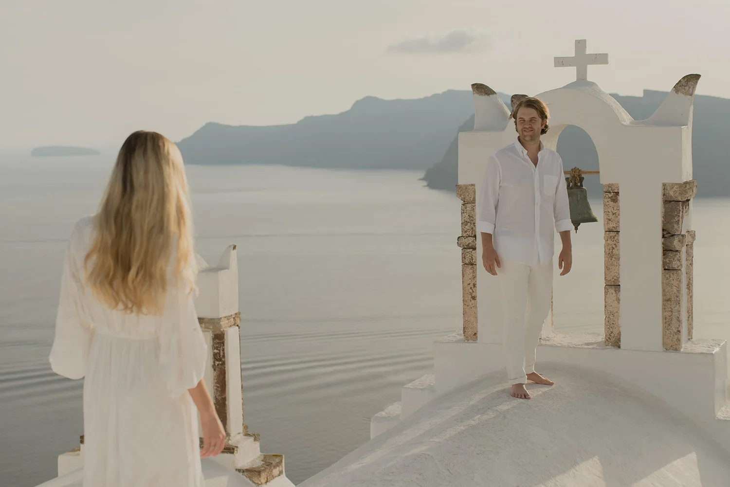 A man and a woman dressed in white are on a white staircase leading to a small chapel with a cross, overlooking the sea and distant mountains in the background.