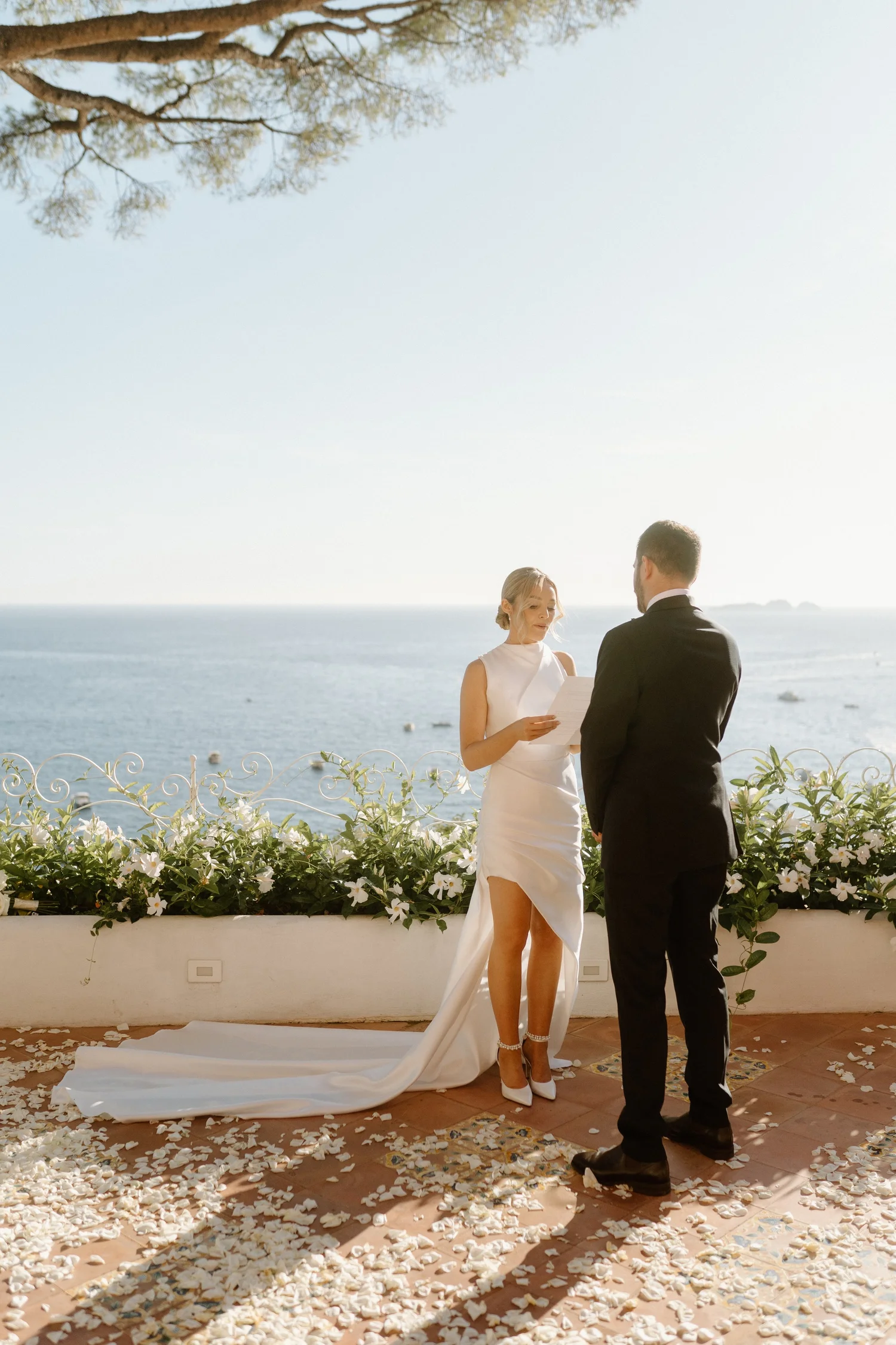 A woman and man stand facing each other during a wedding ceremony outdoors by the water, with the woman reading from a paper, surrounded by white flowers and petals on the ground, in a scenic seaside setting with a tree and calm ocean in the backgrou