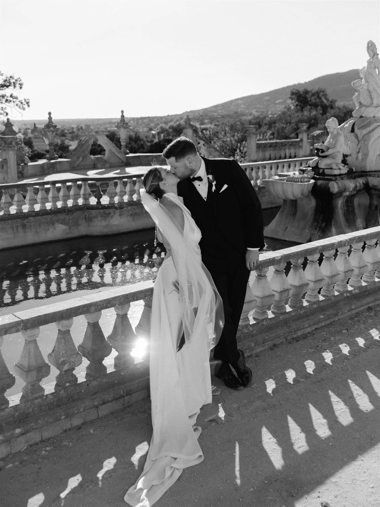 A newlywed couple sharing a kiss on a bridge with ornate balustrades and sculptures, with a scenic mountain and clear sky in the background.