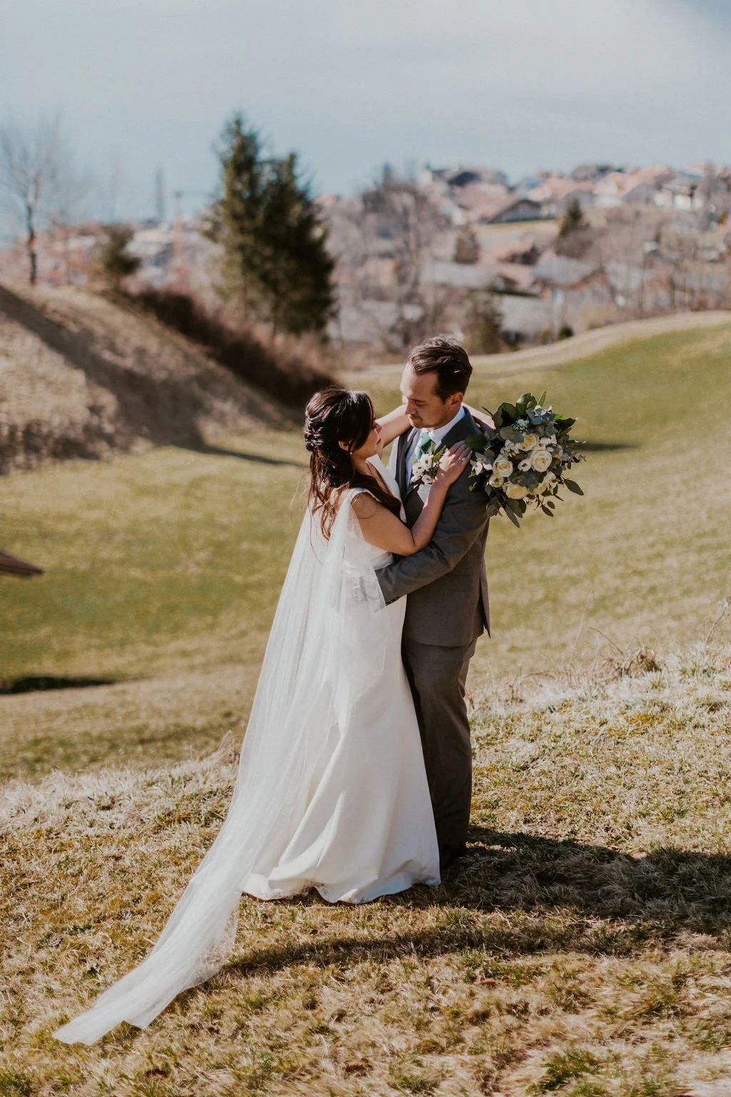 A bride and groom stand close together outdoors during daytime, gazing into each other's eyes on a grassy hill with a scenic background of trees and distant houses.