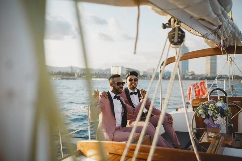 Two men in pink suits with bow ties sitting on a boat during the day, smiling at the camera with a city skyline and water in the background, decorated with flowers.