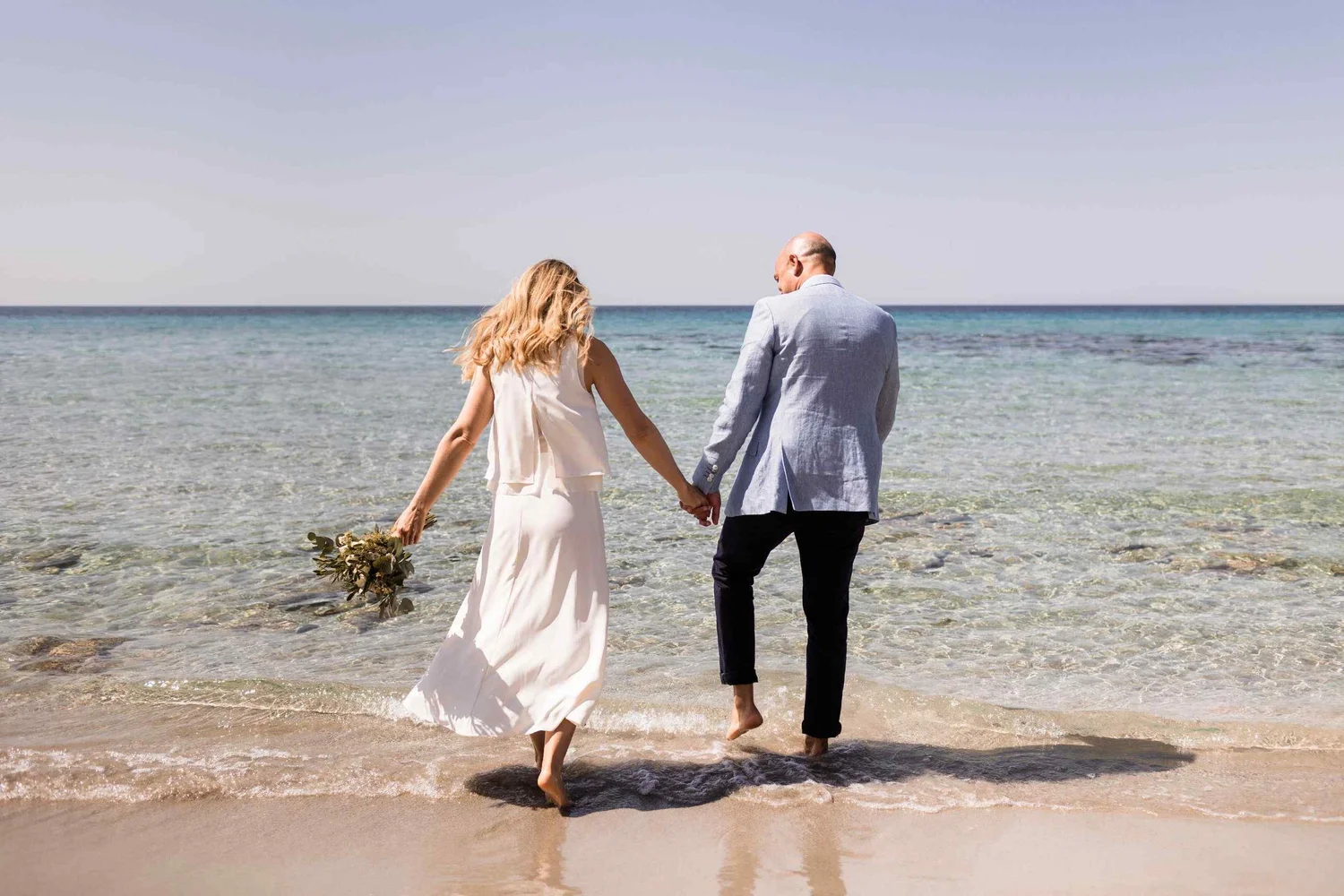 A couple dressed in wedding attire walking hand in hand into the ocean at the beach with clear water and a light blue sky.