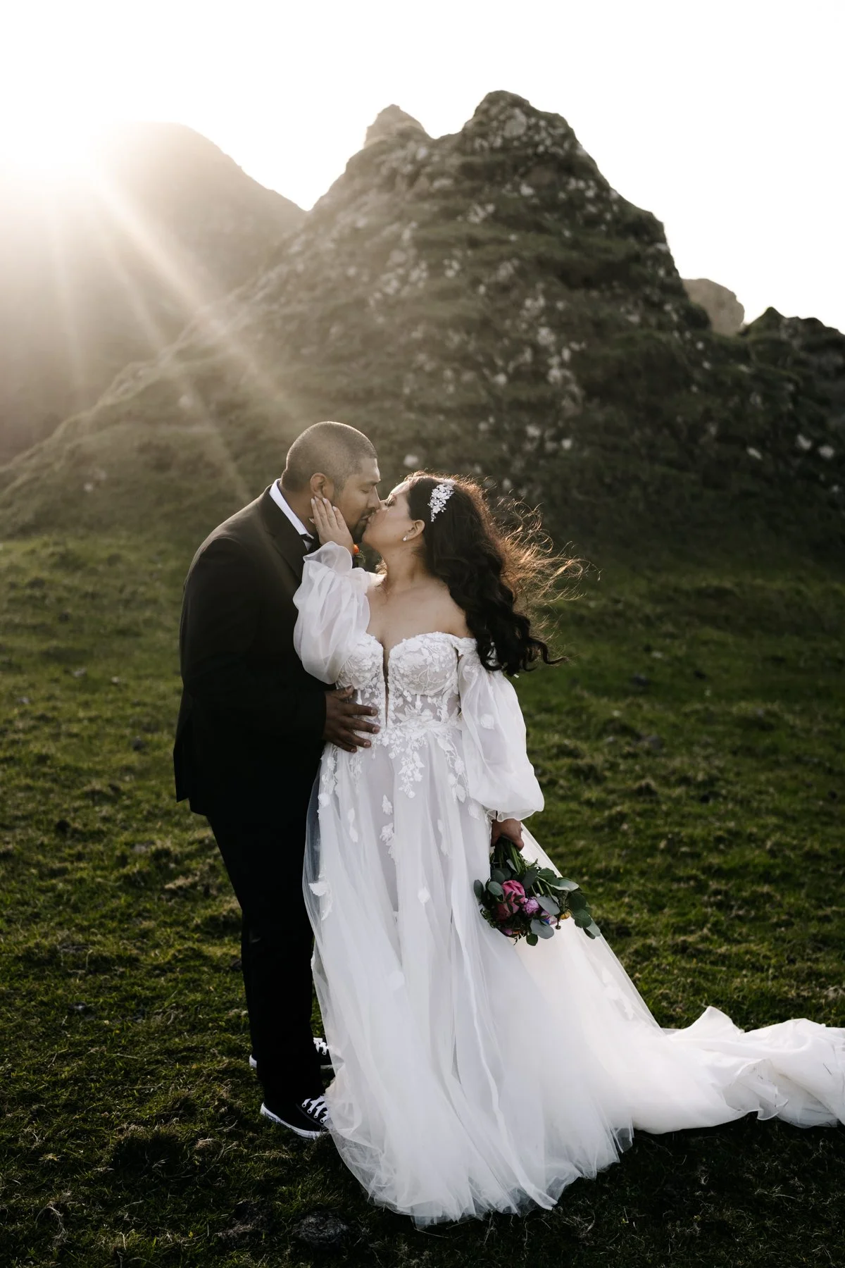 A bride and groom sharing a kiss outdoors during sunset, with a mountainous landscape in the background. The bride is wearing a white wedding gown and holding a bouquet of flowers, while the groom is dressed in a black suit.